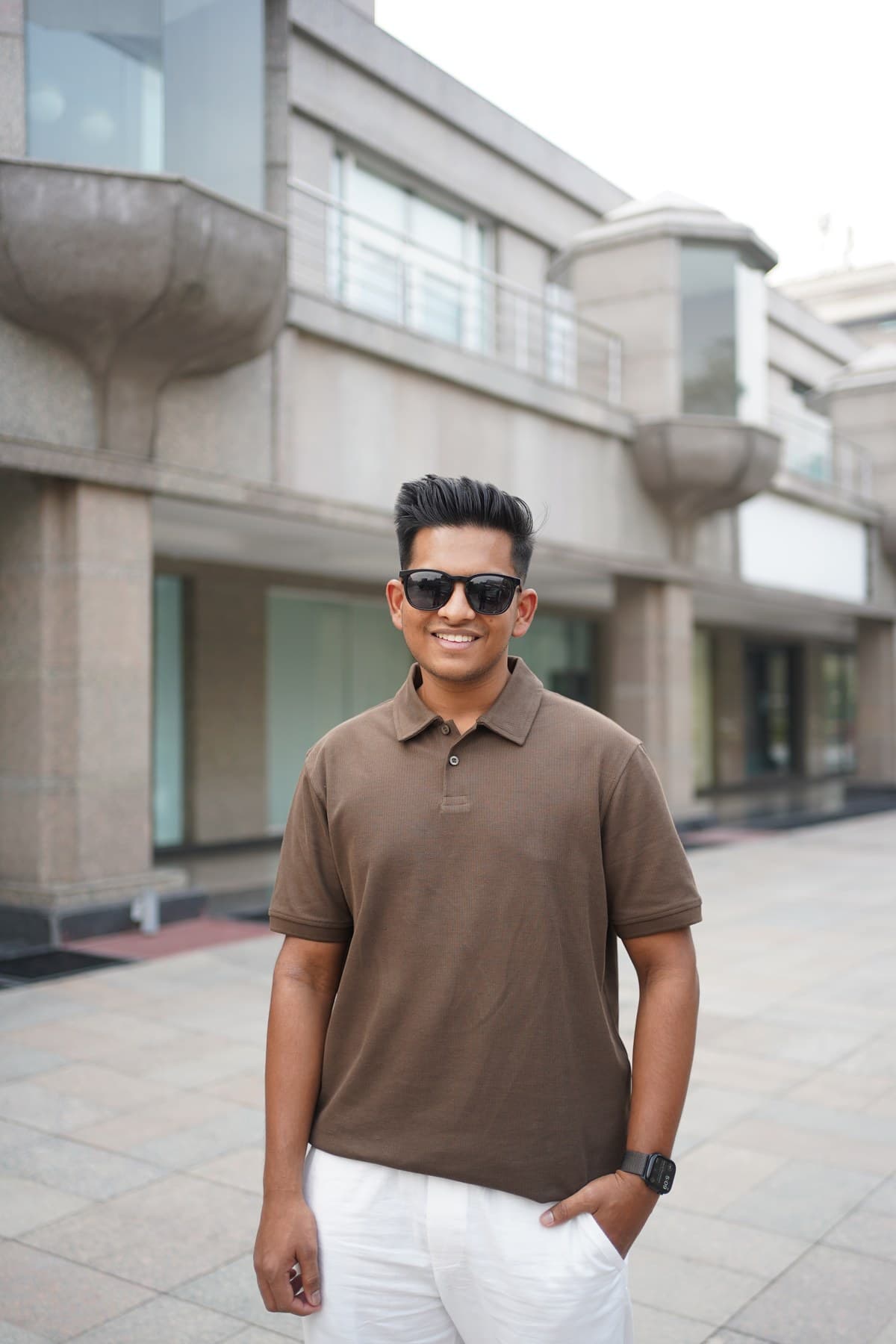 Suhas outside a pale concrete building in bright daylight, in a brown polo and sunglasses, hands in pockets, grinning